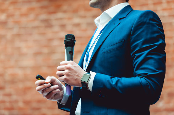 Business and speech topic: Man in a blue suit holding a gray microphone a on a orange bricks background